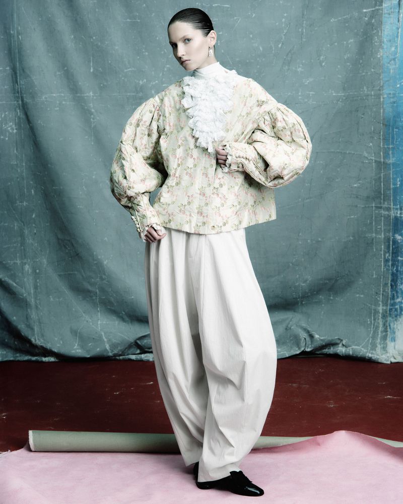 Elegant fashion portrait of a woman in a floral jacket, oversized trousers, and black flats, posing against a textured studio backdrop.