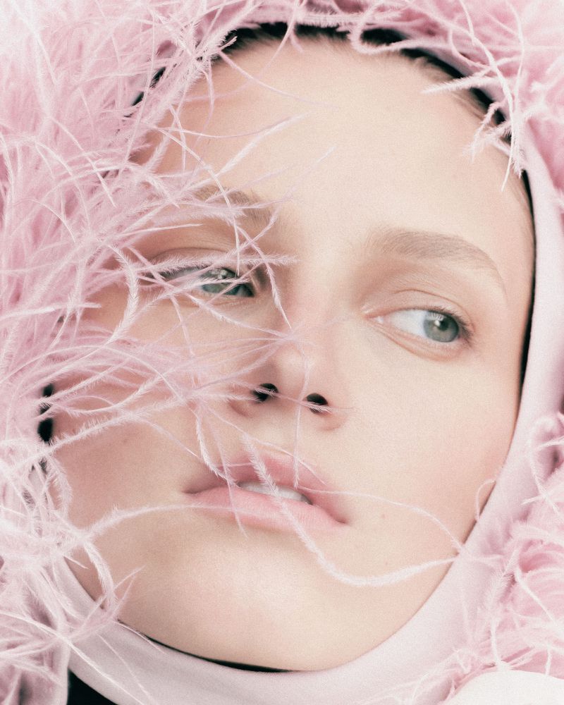 Close-up of a fair-skinned adult female model with neutral facial expression. She has green eyes and is framed by a pink feather headpiece.