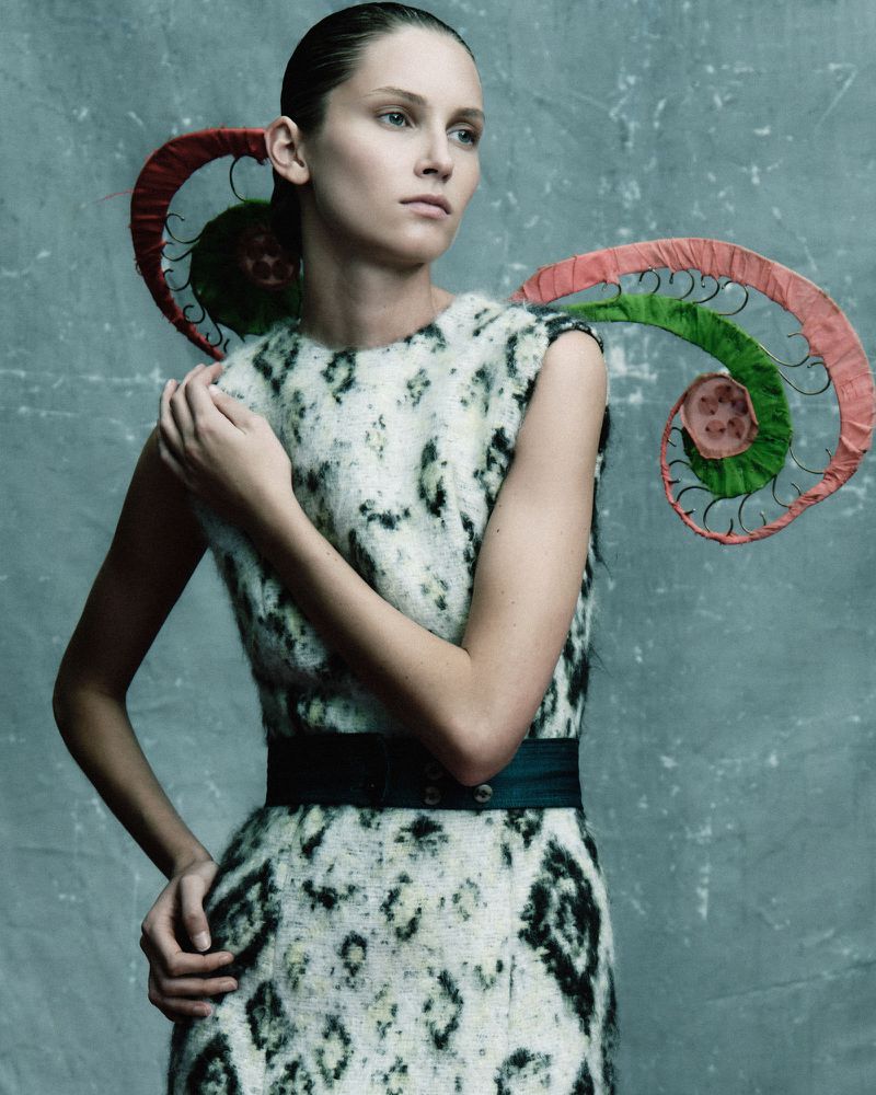 Fashion editorial shot of a female model in a textured dress with artistic spiral decorations against a mottled blue-grey backdrop.