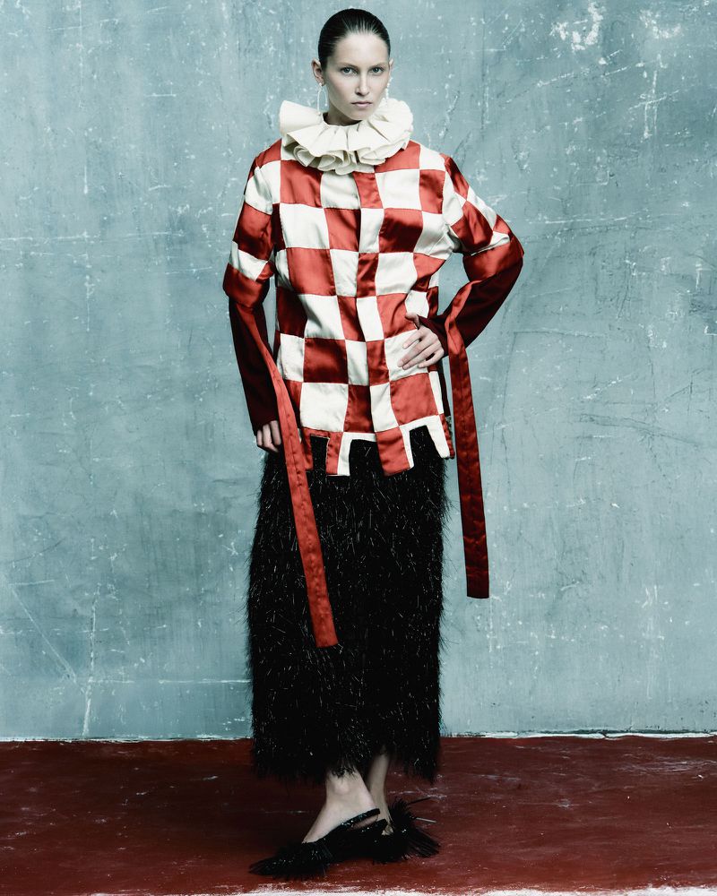 Full-length fashion portrait of a woman wearing a red and white checkered top, black skirt, and black shoes, standing against a textured wall.