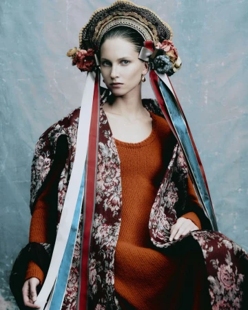 Portrait of a woman in an orange dress and floral coat wearing a traditional headpiece with ribbons against a textured blue backdrop.