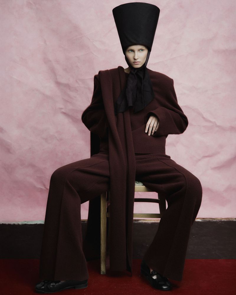 Fashion shot of a model in a burgundy outfit, posed while sitting on a chair against a pink background, wearing a tall black hat.