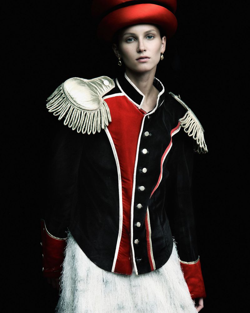 Fashion portrait of a woman in a red hat and ornate black and red jacket with white epaulettes against a dark background.