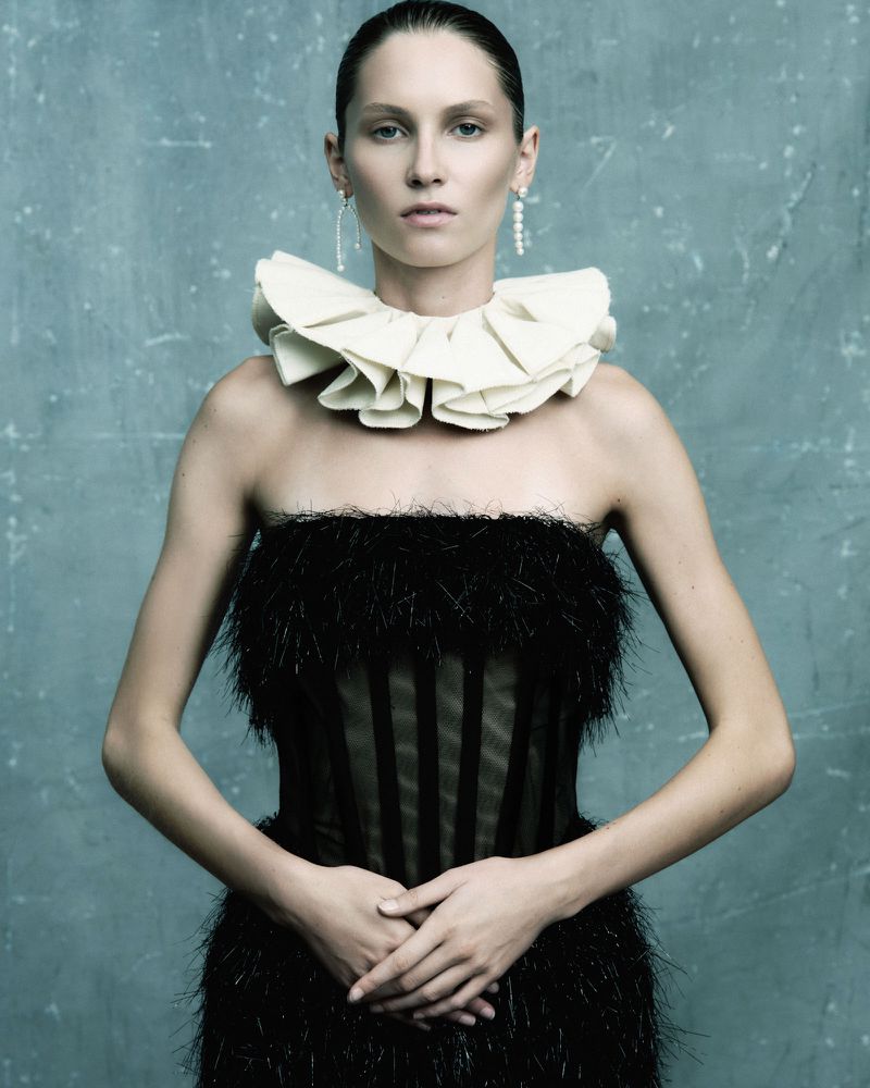 A woman with fair skin poses in a black strapless dress with a ruffled white collar, hands clasped at her waist against a blue background.