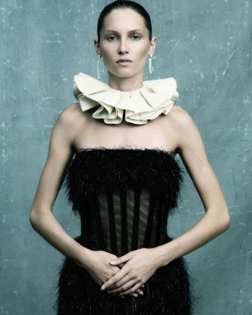 A woman with fair skin poses in a black strapless dress with a ruffled white collar, hands clasped at her waist against a blue background.