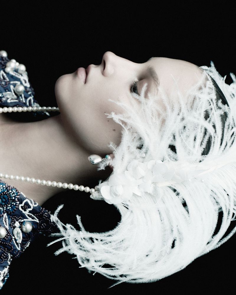 Portrait of a fair-skinned adult female looking upward, adorned with pearl jewelry and a feather headdress against a dark background.