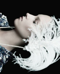 Close-up profile portrait of a woman wearing a feathered headpiece and pearl necklace against a black background.