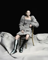 Model seated on a wooden chair wearing a textured grey dress and metallic silver boots against a black backdrop