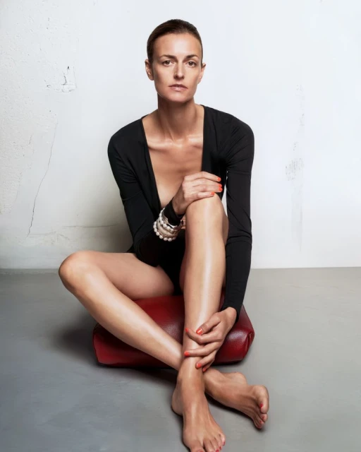 Adult woman in a black bodysuit poses seated on a red cushion indoors with a simple white wall backdrop and grey flooring.