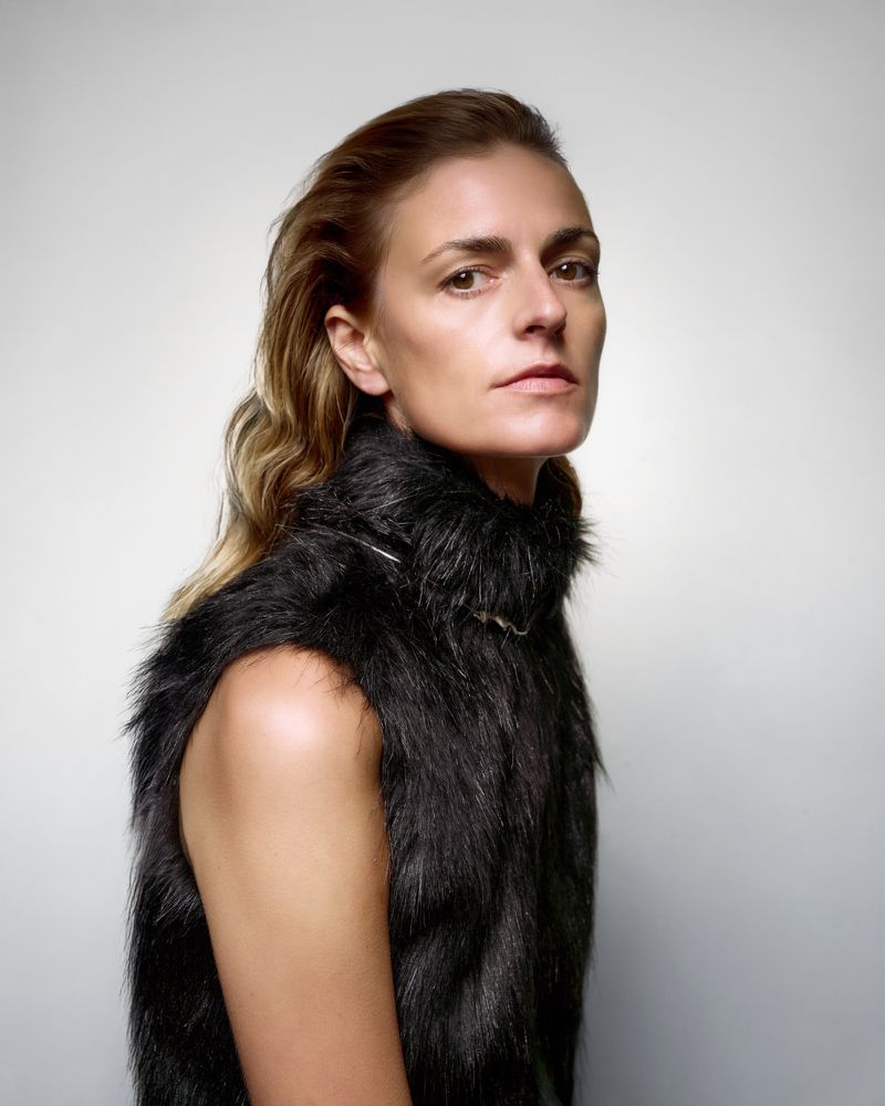 Portrait of an adult female with shoulder-length blonde hair posing in a fur vest against a white background in a photo studio.