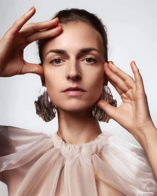 Close-up portrait of an adult woman with hands framing her face, wearing statement earrings and a light pink, translucent top. 