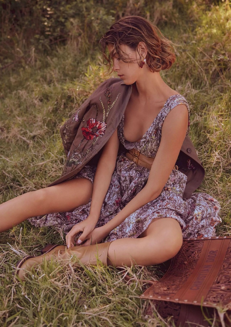 Young woman in a floral dress, jacket and boots sitting in a grassy field with a book, fashion, outdoors, tan boots, neutral lighting.