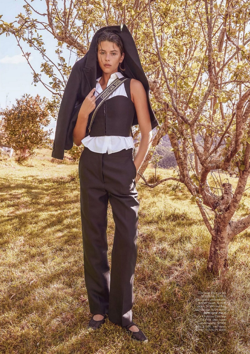 A woman in a black and white outfit and black jacket poses outdoors near trees and tall grass in a fashion shot.