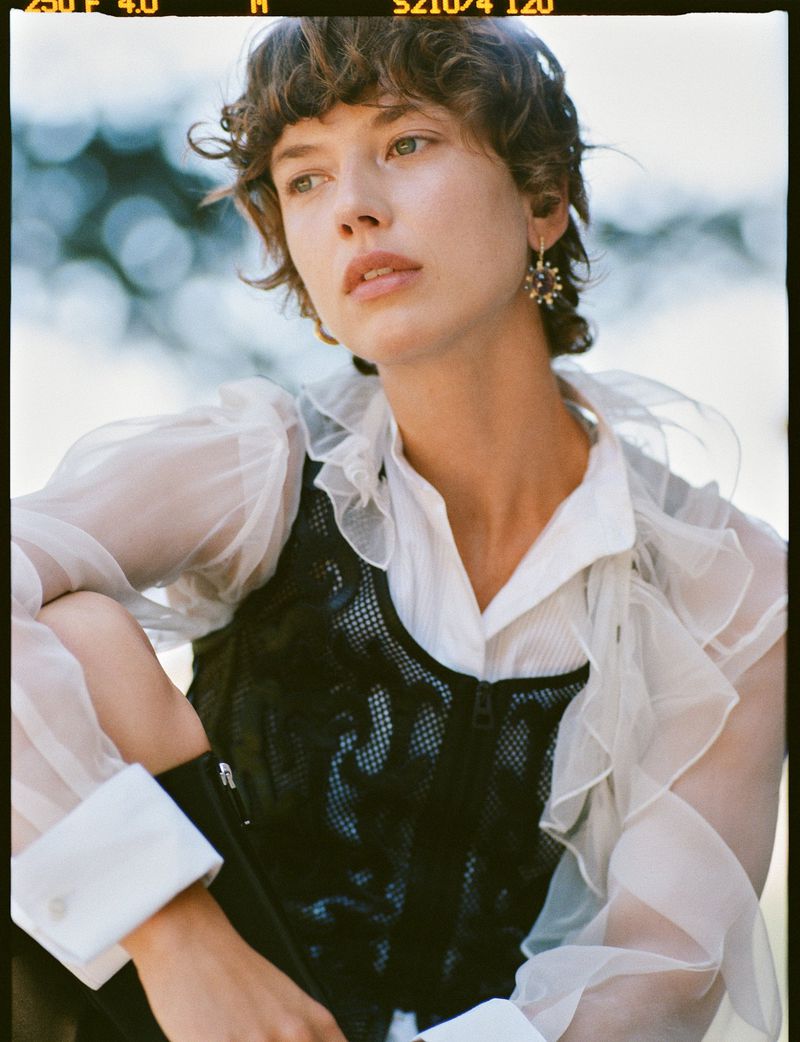 Woman in sheer white ruffled top and black mesh corset outdoors with blurred green background in medium shot, wearing drop earrings.