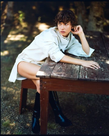 A fashionable woman in a white dress and black boots sits at a rustic wooden table outside in soft sunlight. Fashion, beauty, and outdoor setting.