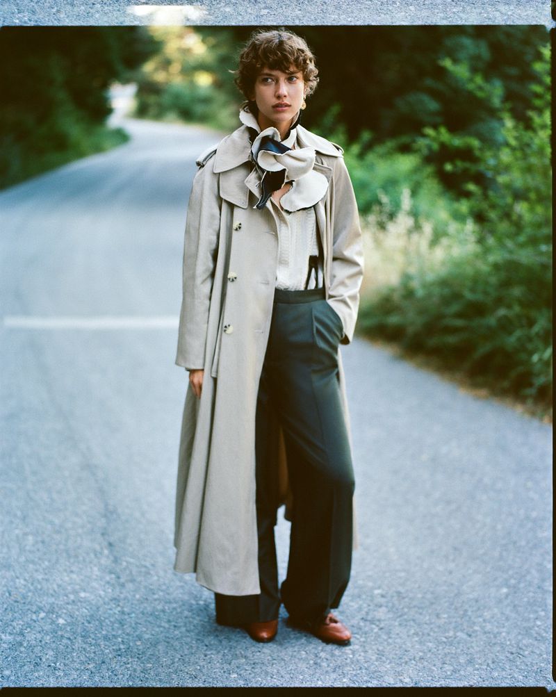 A woman in a trench coat stands on an empty road surrounded by trees. She wears wide-legged pants, a white shirt, and brown shoes.