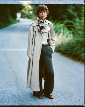 A woman in a trench coat stands on an empty road surrounded by trees. She wears wide-legged pants, a white shirt, and brown shoes.