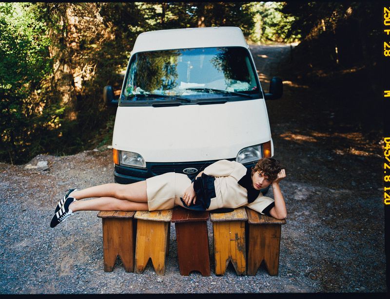 A young adult lies on a row of stools in front of a white Ford van on a gravel road surrounded by trees in natural light.