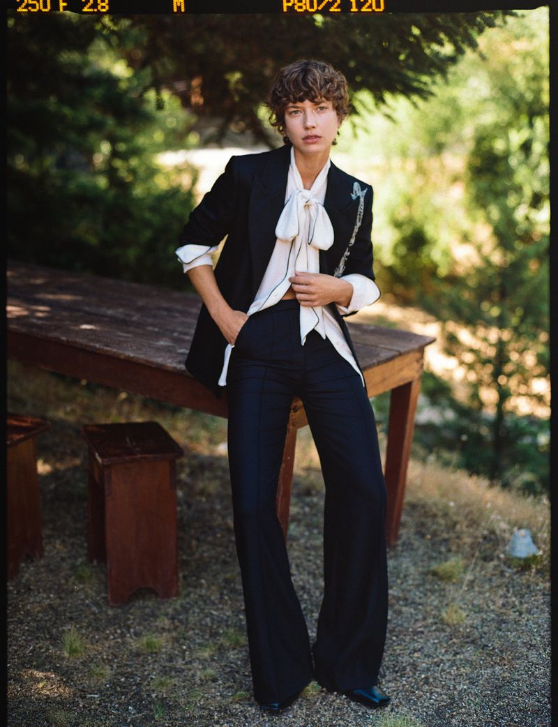 A slender woman poses outdoors wearing a black blazer, white blouse with a bow, dark trousers, and black shoes, with trees in the background.
