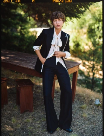 A slender woman poses outdoors wearing a black blazer, white blouse with a bow, dark trousers, and black shoes, with trees in the background.