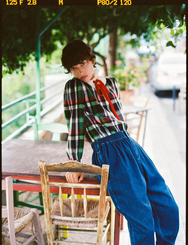Adult female posing outdoors in a plaid top and blue pants, leaning against a table with chairs on a stone terrace.