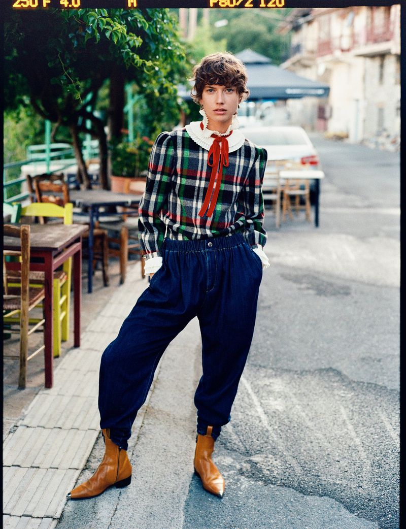 Woman posing in a plaid top, blue pants, and boots on a cobblestone street with an outdoor cafe and vehicle in the background.