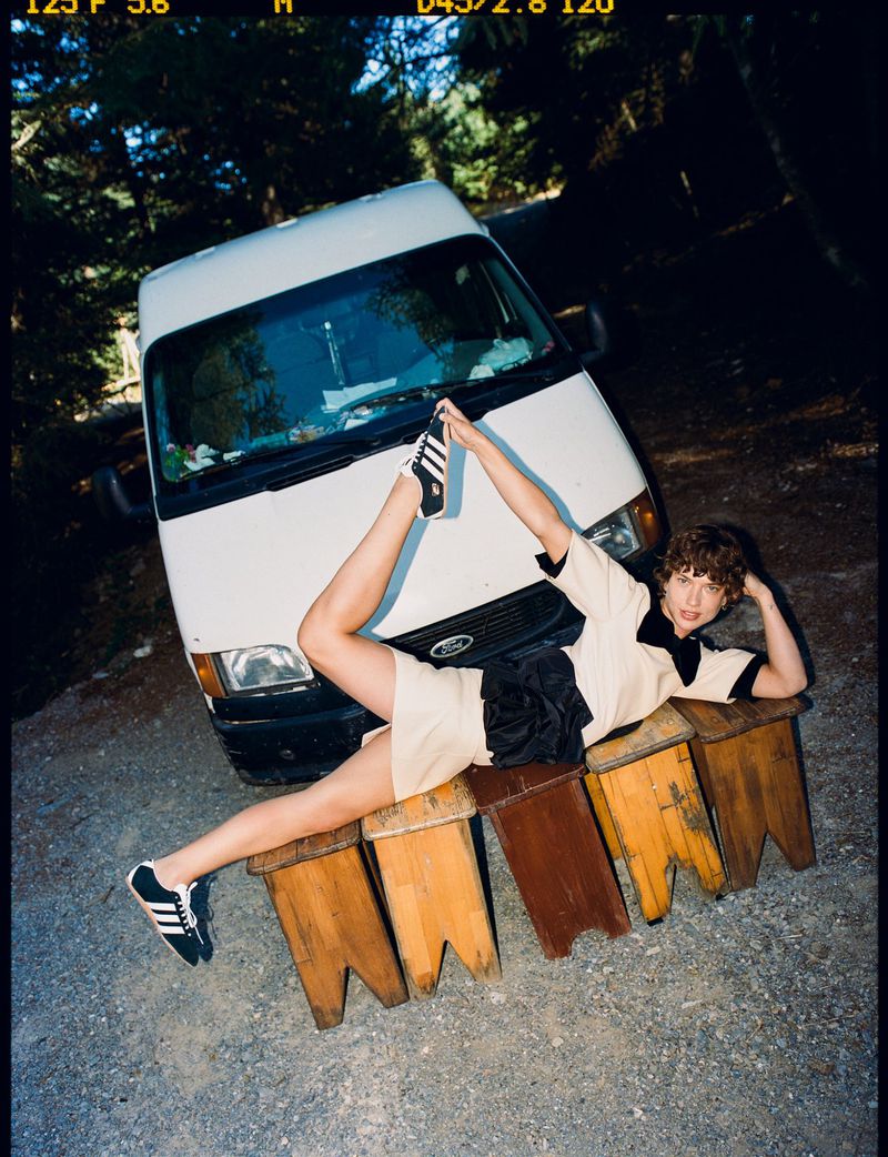 A young adult lying on wooden benches, posing with legs raised against a backdrop of a white van and trees, captured in outdoor lighting.