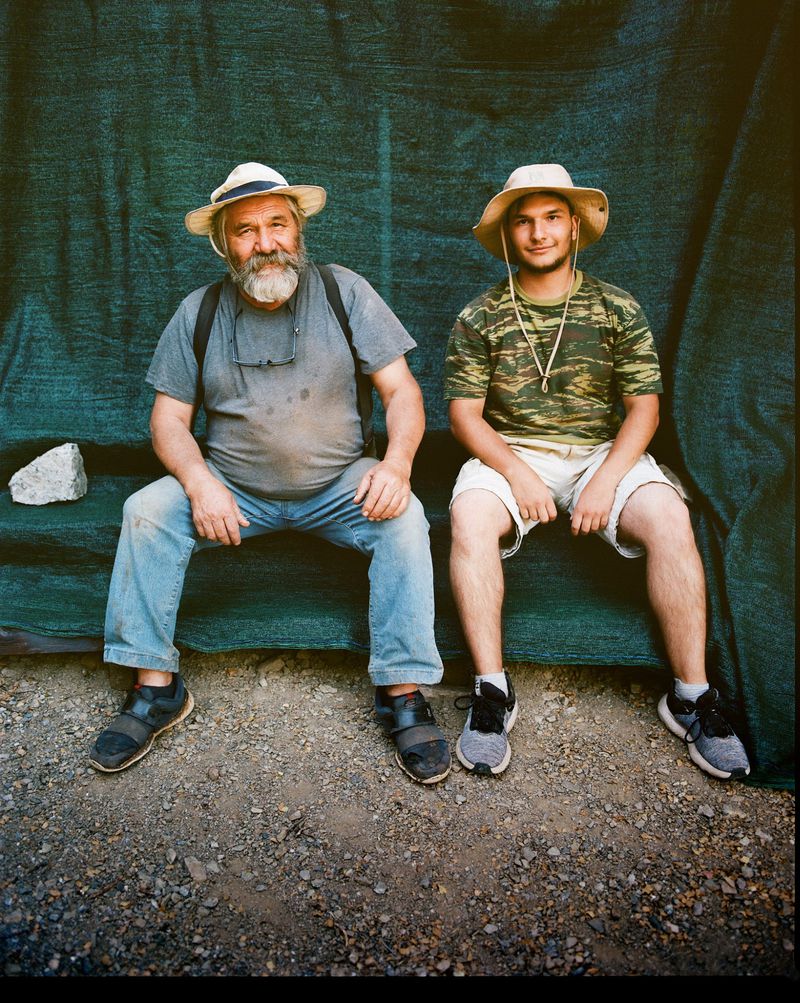 Outdoor portrait of an older and younger man wearing hats, seated together, posing in front of a green fabric backdrop in natural light.