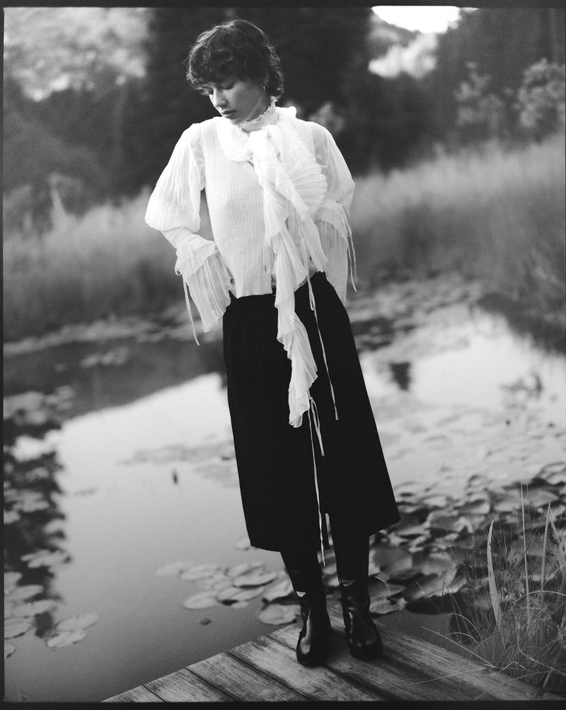 Woman in ruffled blouse and dark skirt stands on wooden dock by water with tall grass and trees in background in black and white.