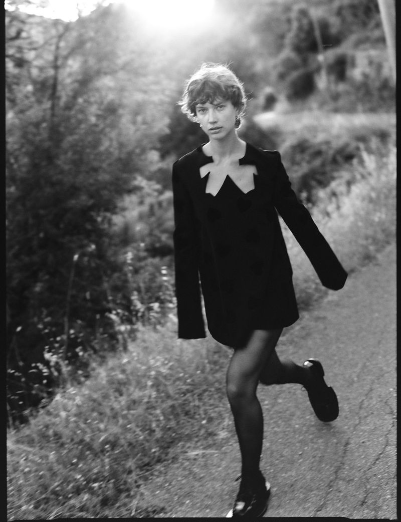 A young adult female model in a black dress and tights running on a rural road with trees, photographed in black and white.