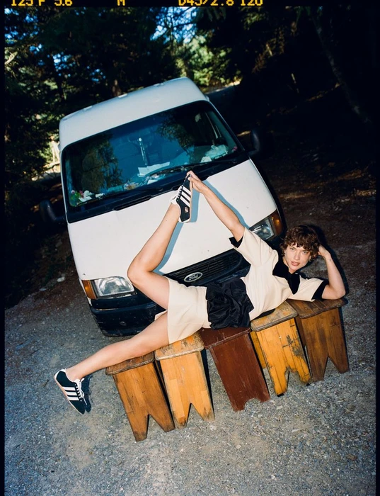 Model posing in cream outfit and sneakers stretched across wooden stools in front of a white van