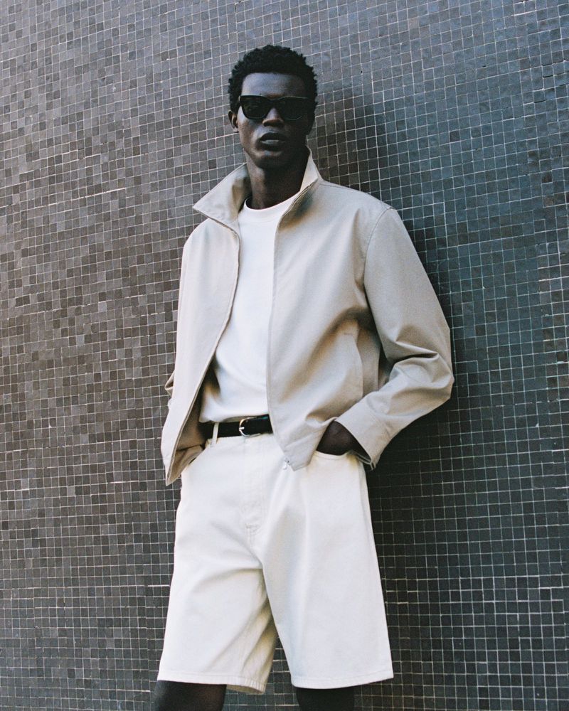 A dark-skinned male model poses against a grey tile wall wearing sunglasses, a beige jacket, a white t-shirt, and white shorts with his hands in his pockets.