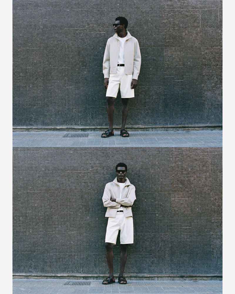 A Black male model stands in front of a textured wall, wearing sunglasses, an open beige jacket, white t-shirt, cream shorts, and sandals.