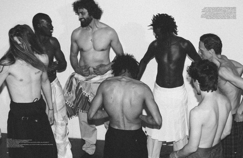 Black and white image featuring a group of bare-chested men in a circle, wearing skirts or pants, against a light backdrop.