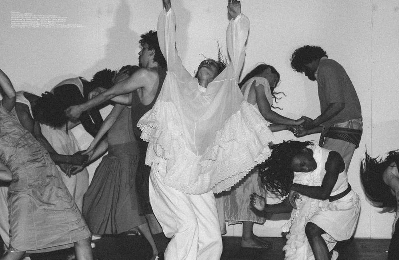 Group of people dancing indoors, arms interlocked, wearing a variety of dresses and tops in a black and white editorial photograph.
