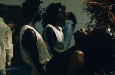 Black man in a white tank top standing before a mirrored wall reflecting multiple figures in a moody, dimly lit interior setting.