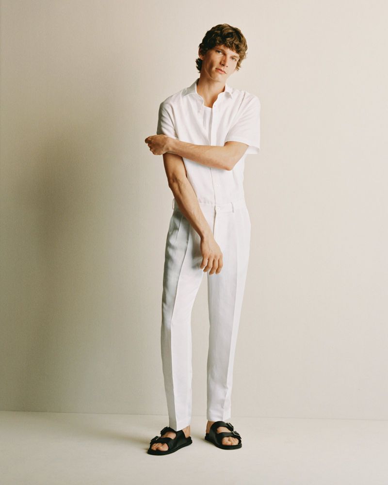 An adult male model with curly brown hair poses in a white button-down shirt, white slacks, and black sandals in a studio setting.