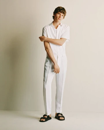 An adult male model with curly brown hair poses in a white button-down shirt, white slacks, and black sandals in a studio setting.