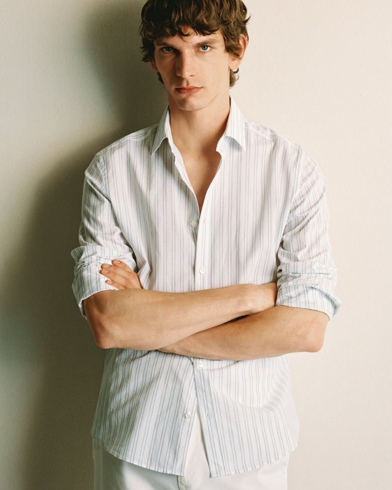 Portrait of a light-skinned adult male in a button-down shirt with arms crossed, posing indoors against an off-white wall in a fashion setting.