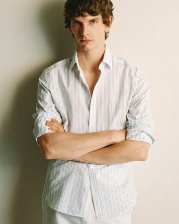 Portrait of a light-skinned adult male in a button-down shirt with arms crossed, posing indoors against an off-white wall in a fashion setting.