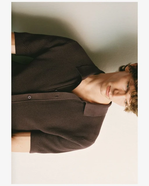 Portrait of a young adult man wearing a brown knit polo shirt, posing indoors with a subtle smile against a neutral background.