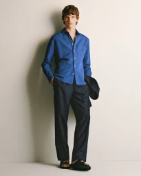 Full-length portrait of a male model wearing a blue button-down shirt and dark trousers against a neutral white wall.