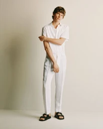 Man wearing a white short-sleeve shirt, white trousers, and black sandals against a neutral studio background.