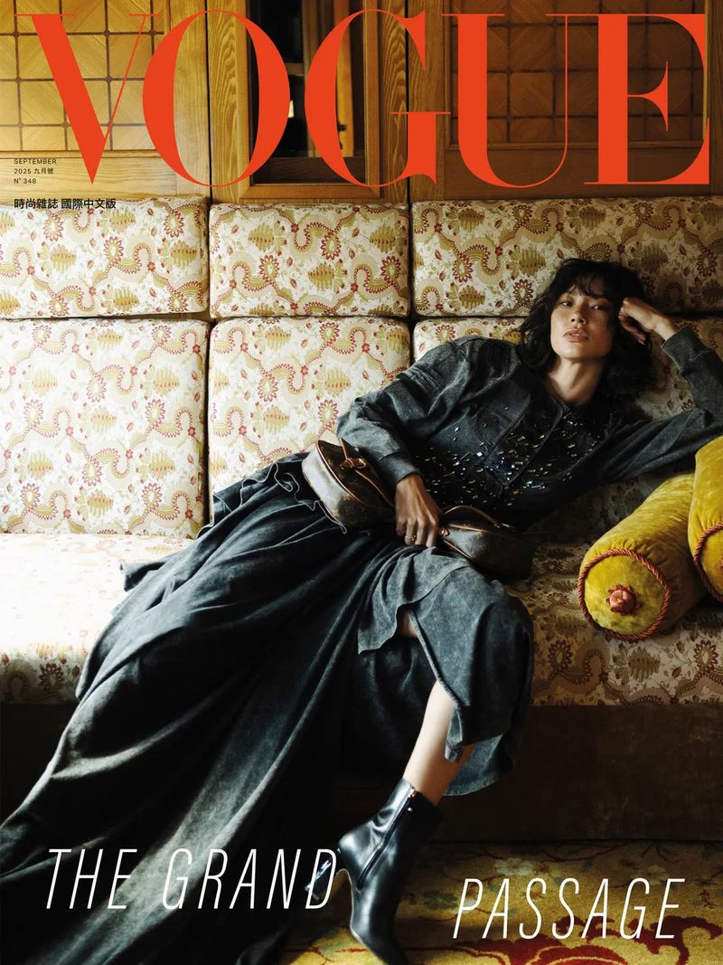Vogue cover featuring a woman in a long grey dress and black boots lounging on an ornate patterned couch in an indoor setting.