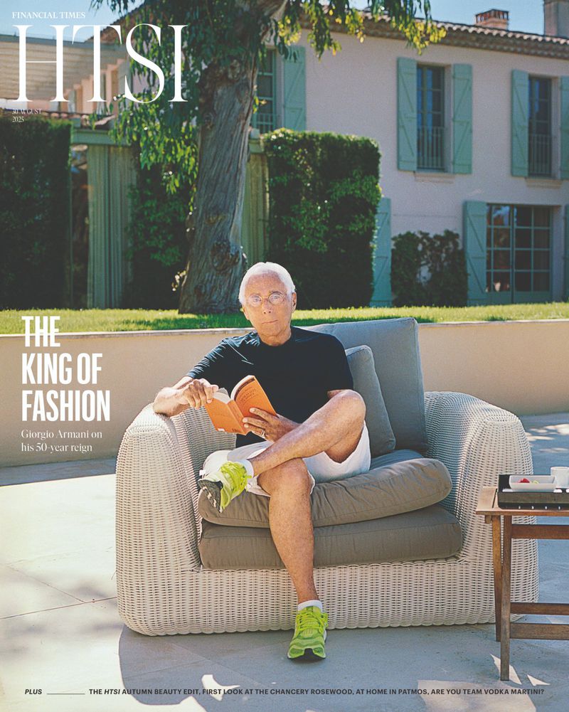 Giorgio Armani sits in an outdoor chair reading a book, cover of HTSI Financial Times, with a house and lawn in the background.