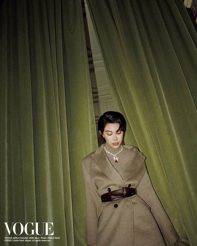 Fashion editorial shot of a brunette woman in a belted coat posing in front of green velvet curtains, illuminated by soft, studio lighting.