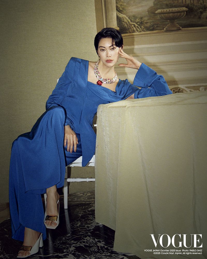 Elegant woman in blue dress and statement necklace poses indoors. Fashion editorial shot with luxurious details, marble floor, and subdued lighting.