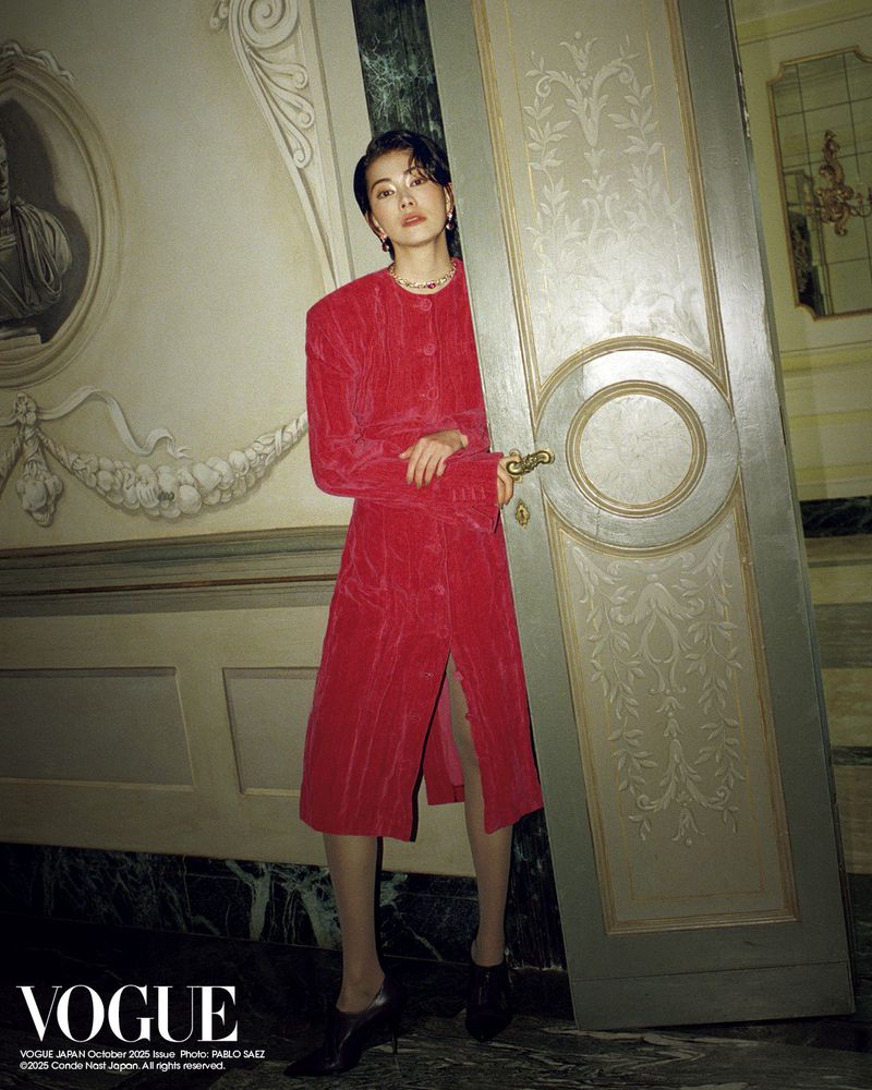 An adult woman stands in a vintage room wearing a red midi dress and black heels, one hand on a large doorway, in an editorial shot.