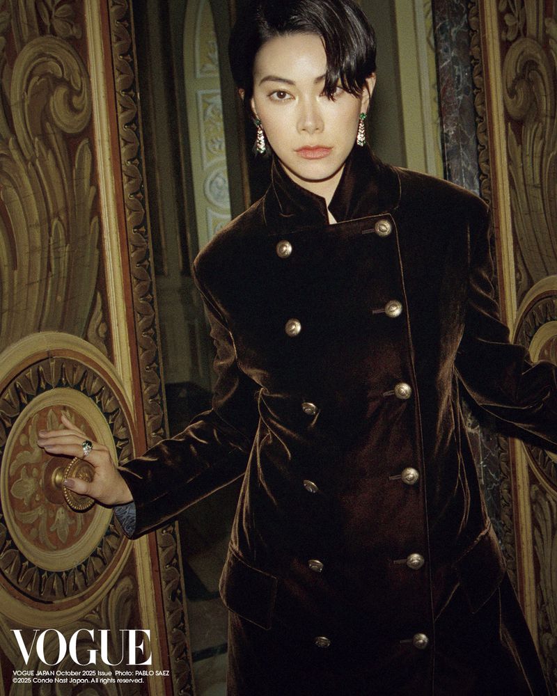 Elegant woman in a double-breasted brown velvet coat opening an ornate gold door in an opulent interior with soft, diffused lighting.