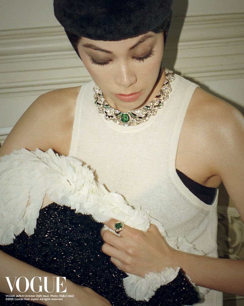 Woman wearing a black cap and white and green jewelry poses indoors with a black and white feathered garment in a studio shot.
